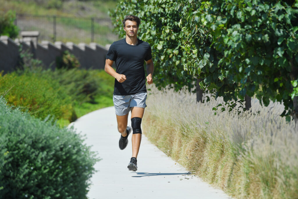 A man running outside in the sun with a knee brace on his left leg. 